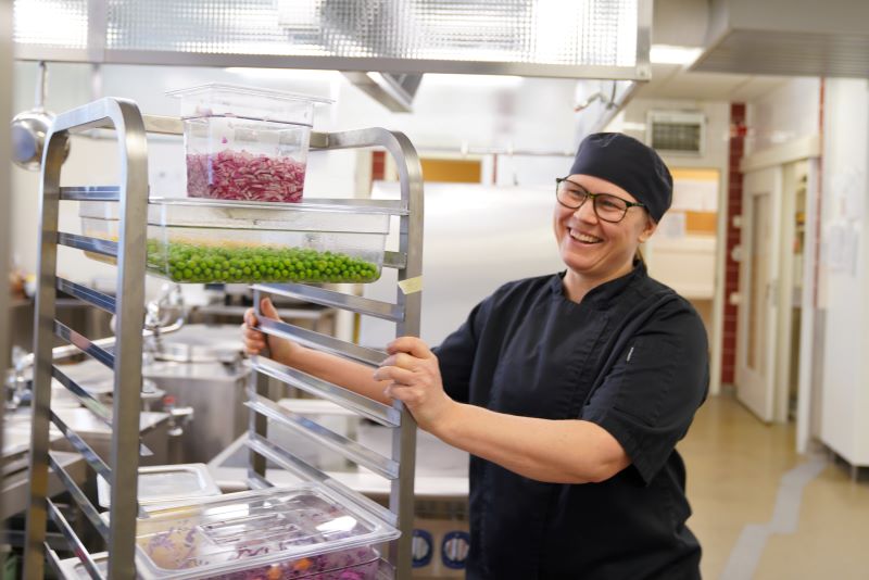 En kvinna i kockkläder med en vagn lastad med grönsaker.